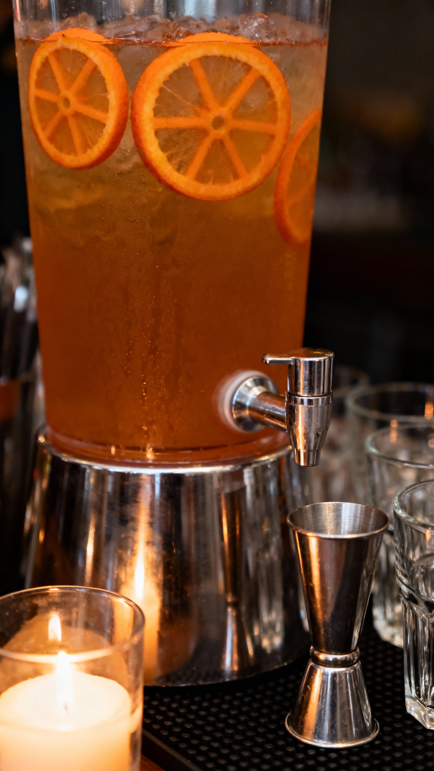 Closeup of batch cocktail dispenser with amber drink, floating orange wheels, glassware nearby, stai